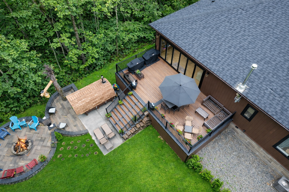 Aerial view of a custom-build home featuring a large deck, landscaped backyard, fire pit area, and forest surrounding 