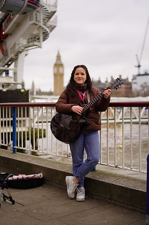A woman stood in front of the London Eye and River Thames, holding a guitar