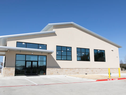 Exterior view of the Rayburn Electric Cooperative recreational facility in Rockwall featuring a light colored building with black framed windows, stone base accents, and a covered entrance. The modern structure sits beneath a clear blue sky with paved parking and landscaped grounds in the foreground.