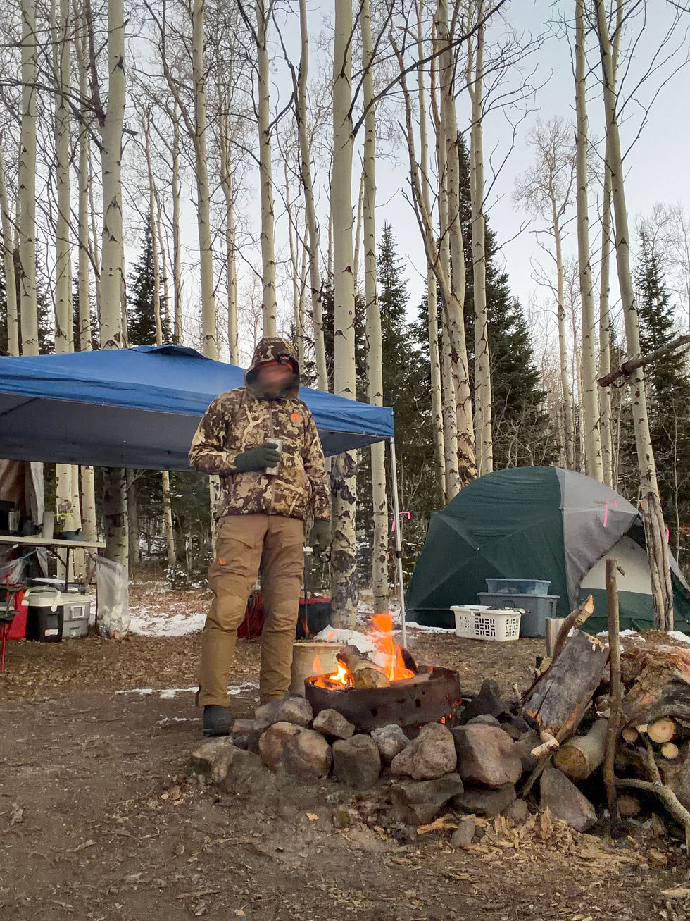 A person in camo gear stands by a campfire holding a mug in a forest with tall trees. A tent and blue canopy are in the background.