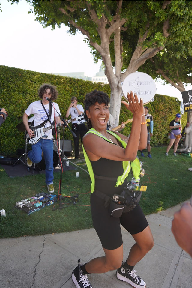 A demonstrator dances to a band in a shaded area. The demonstator, an African American woman, wears a highlighter-yellow traffic vest over a black jumpsuit; around her waist is a holster for a camera. She is captured mid-dance with a tambourine in her hand. The tambourine has writing on its front "Tanjareen's tambourine" in stylized writing in black. Behind her standing in the grass is a male gutarist who plays a black and white electric guitar and sings into a microphone. Some other photographers and picketers are visible. 