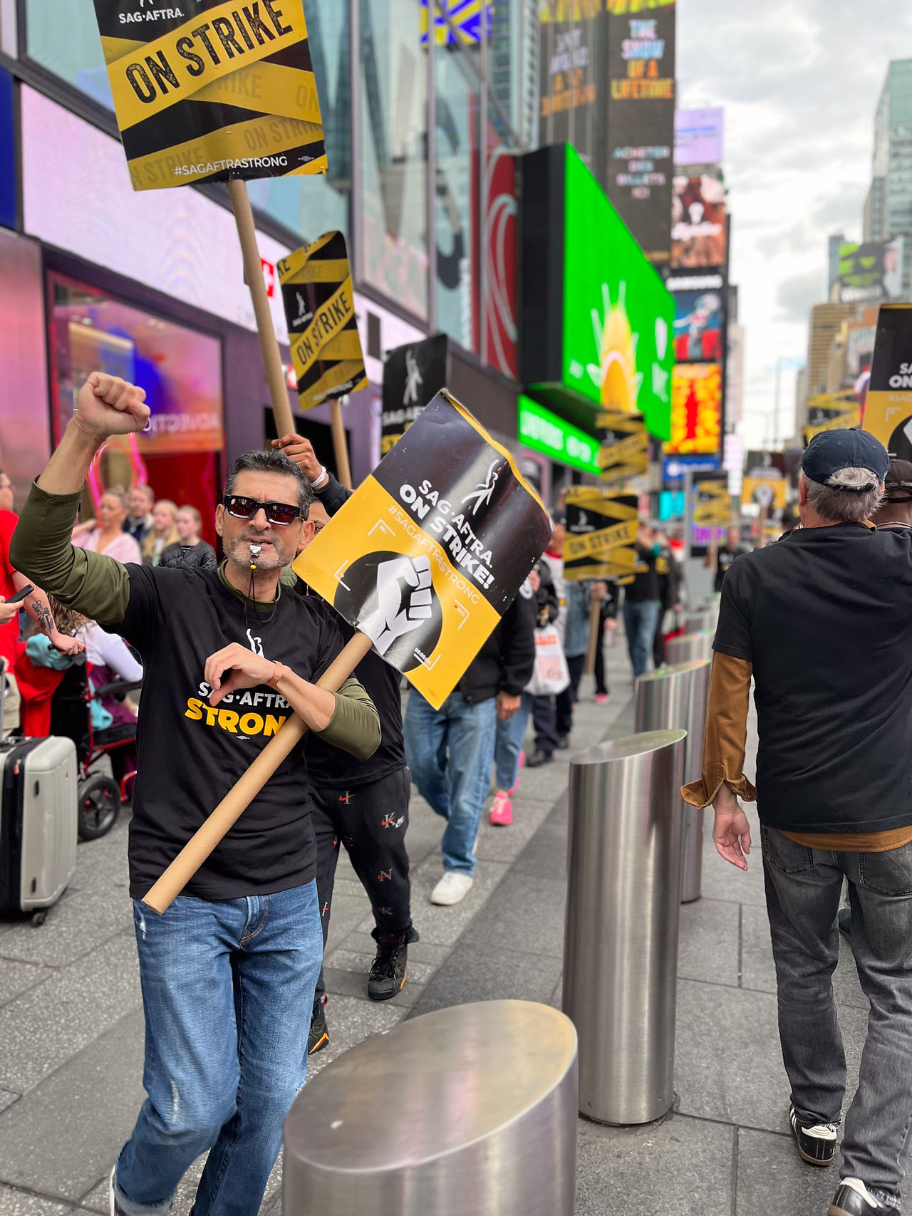 A man walking the line with in Time Square. The man appears on the left of the photo. He wears sunglasses, a black "SAG-AFTRA Strong" t-shirt over a green long-sleeved shirt and jeans. Cradled in his left elbow is a black-and-yellow "SAG-AFTRA On Strike!" graphic picket sign; his right hand is raised in a solidarity fist and there is a silver whistle in his teeth. Other demonstrators are visible both behind the man and on the right of the photo. In the far left background are pedestrians; also visible are billboard signs and city buildings. 