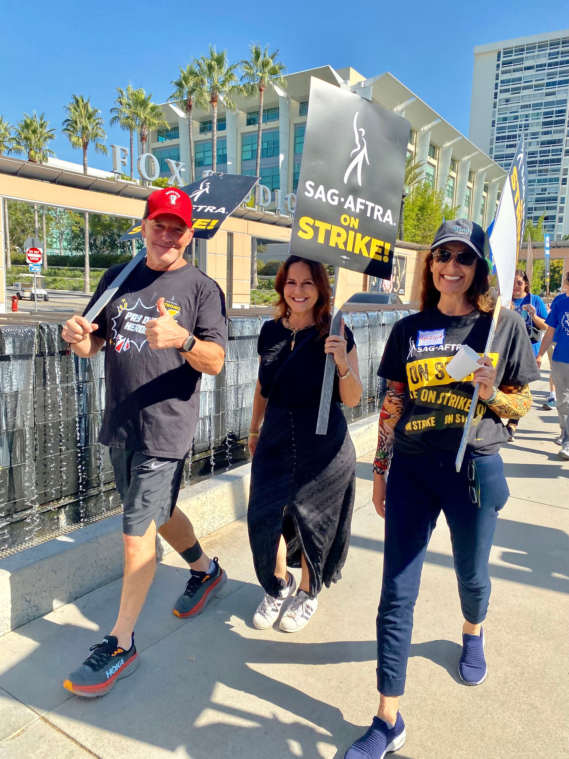 Member Clark Gregg, far left, walking with two other women. The three make a formation of a straight line. Gregg wears a red cap, a black SAG-AFTRA "Pay Our Heroes" t-shirt, and black Nike basketball shorts. He holds a "SAG-AFTRA On Strike!" picket sign in his right hand, and gives a "thumbs up" to the  photographer with his left hand. The woman in the center is a shorter height and wears a black shirt and a dark-colored skirt. She holds a "SAG-AFTRA On Strike!" picket sign and smiles toward the photographer. The woman on the far right wears a dark ball cap and sunglasses, a multicolored shirt with an unknown pattern under her black "SAG-AFTRA On Strike!" graphic t-shirt and blue jeans. She holds a picket sign and smiles at the camera. The Fox Studios sign, palm trees, water fixture and buildings are visible in the background. 