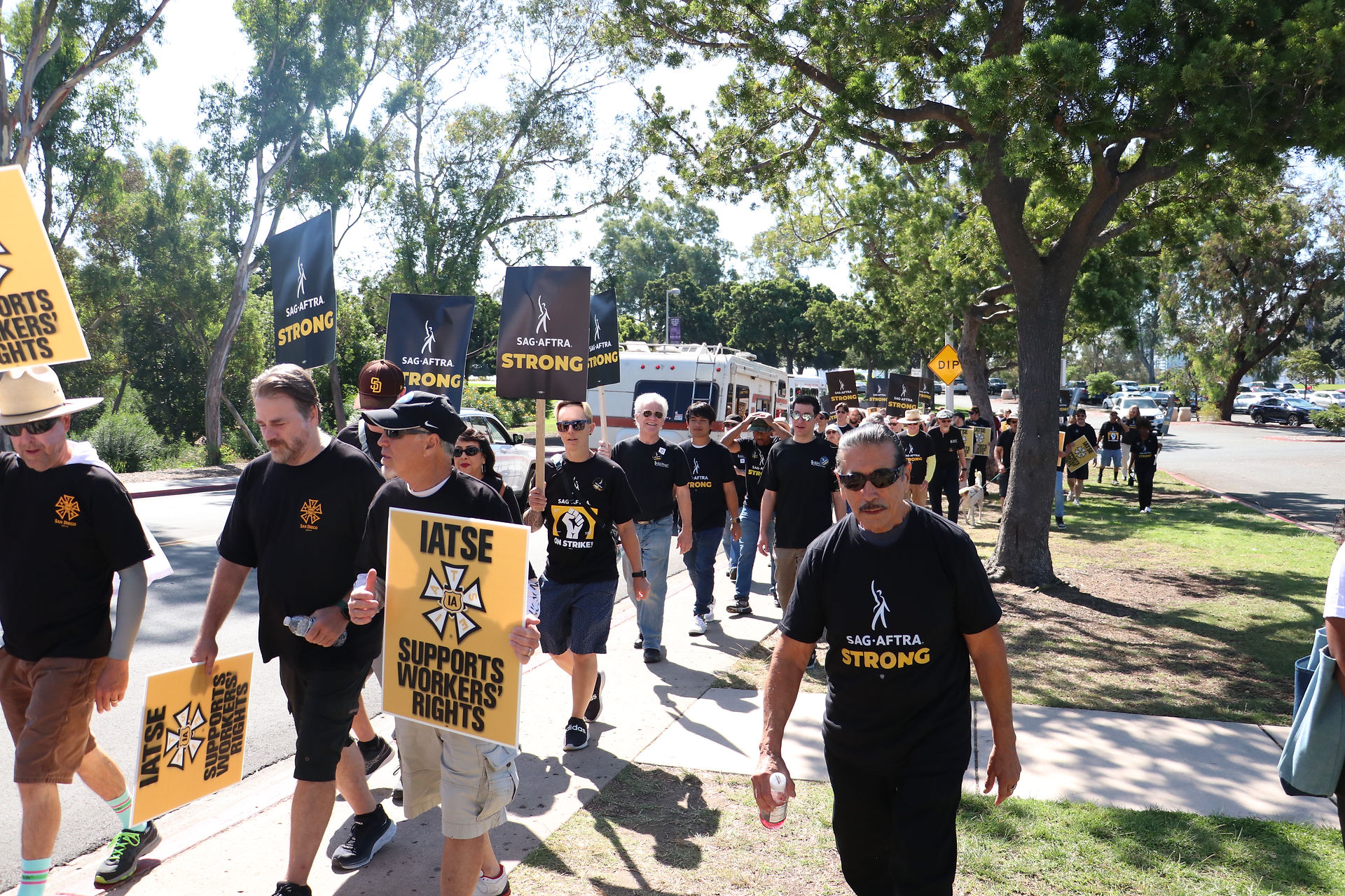 A candid shot of San Diego Local members walking down a street. The members walk in a procession, far left; many wear black SAG-AFTRA and IATSE t-shirts. SAG-AFTRA and IATSE picket signs seen throughout; also visible are trees and cars within a parking lot.