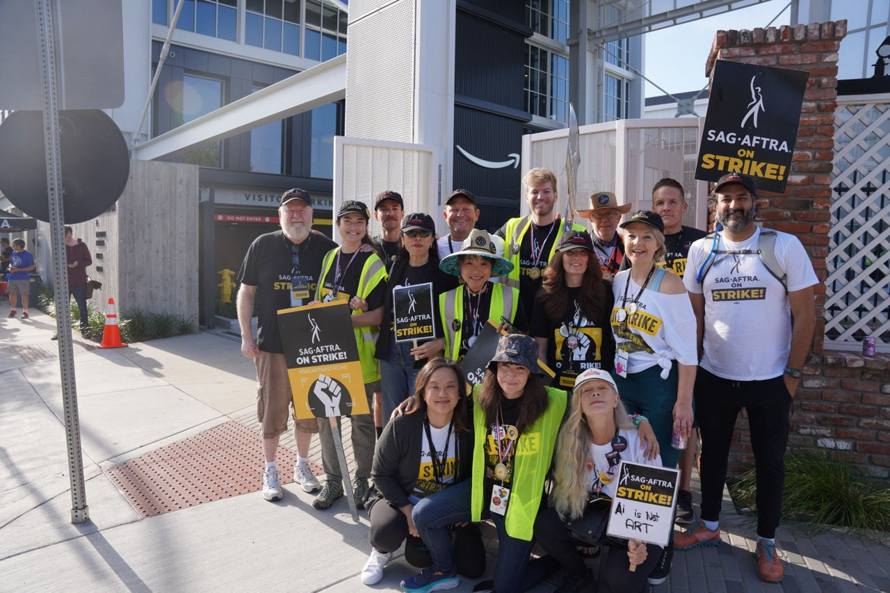 A group photo of the TV/Theatrical/Streaming Negotiation Committee members, staff, and SAG-AFTRA Strike team captains. The group is arranged in a few rows with those in front kneeling on the ground. Among those visible are LA Local President Jodi Long, committee members Frances Fischer, Kevin West and Woody Schultz. Many in the crowd wear SAG-AFTRA Strike t-shirts; captains wear traffic vest over their shirts. Signs seen throughout. Behind them is the gate to a parking lot and a large building. 