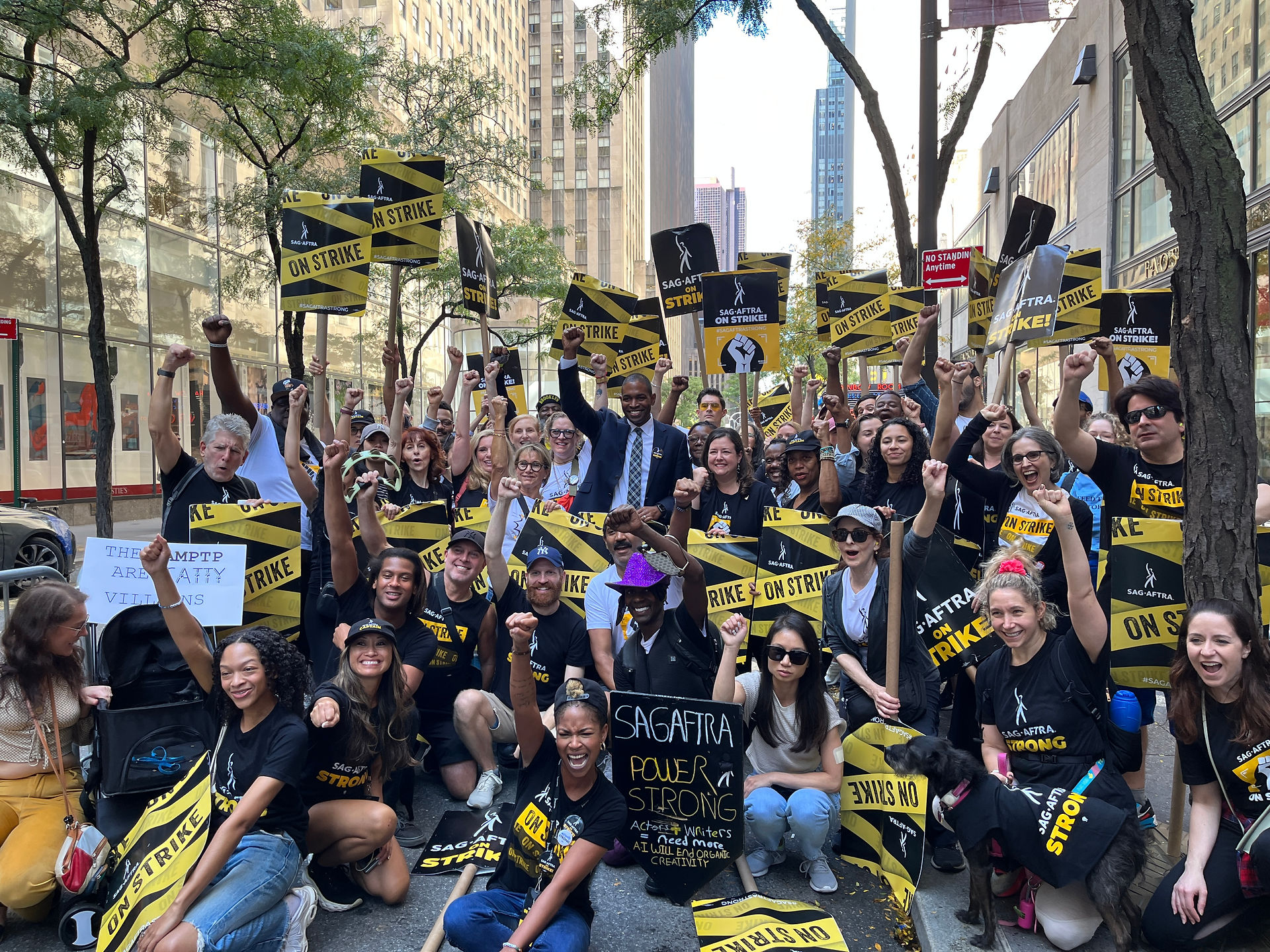 A group photo of New York Local members with Lt. Governor Antonio Delgado, an Afro-Latino male who stands in the middle of the diverse crowd in a dark suit blazer, shirt and tie and "SAG-AFTRA On Strike!" button with his arms raised. The members, who are arranged in a few rows, with those in the front crouching or otherwise sitting on the ground follow suite and raise their fists. Many wear SAG-AFTRA Strike t-shirts. Picket signs seen throughout. In the background are scenes of a street: buildings and trees. 
