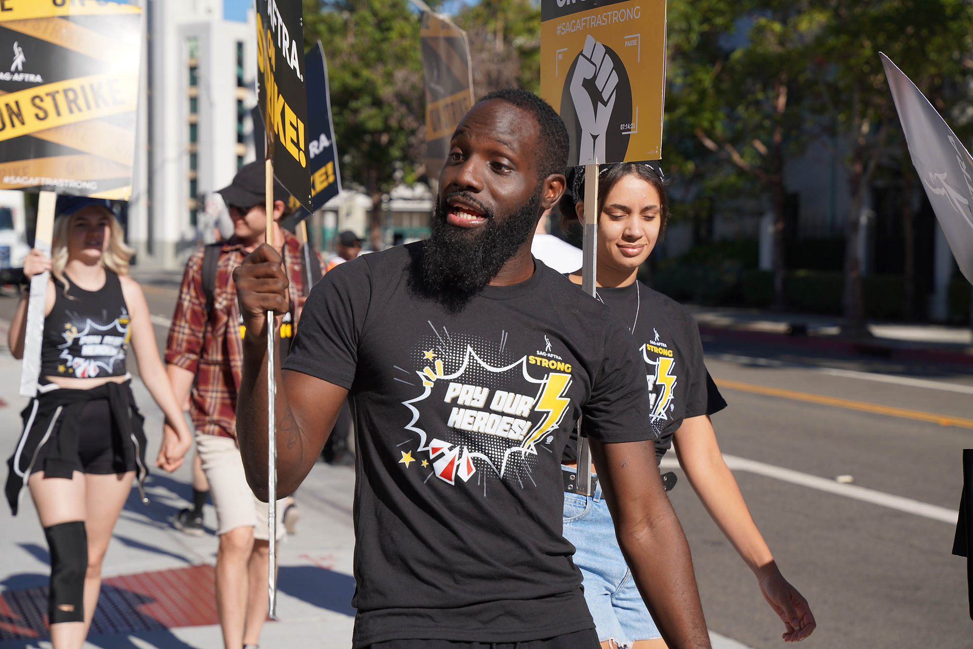 An African American man with a beard walks the picket line. He stands in the foreground of the photo; he wears a black "Pay Our Heroes" t-shirt and holds a "SAG-AFTRA On Strike!" picket sign. Behind him are other walkers as well as an empty street and white buildings on the other side of the road. 
