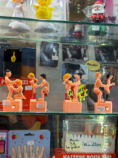 Erotic novelty souvenirs and provocative magnets in a Paris gift shop window, featuring humorous adult-themed figurines and vintage-style postcards.