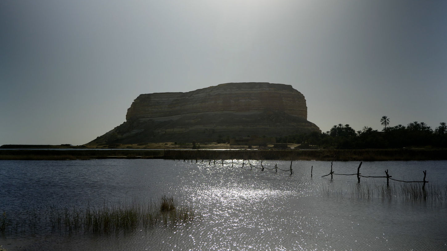 A lake in front of a sandstone hill and trees at the Siwa Oasis in the Sauna Channel episode: Three Springs