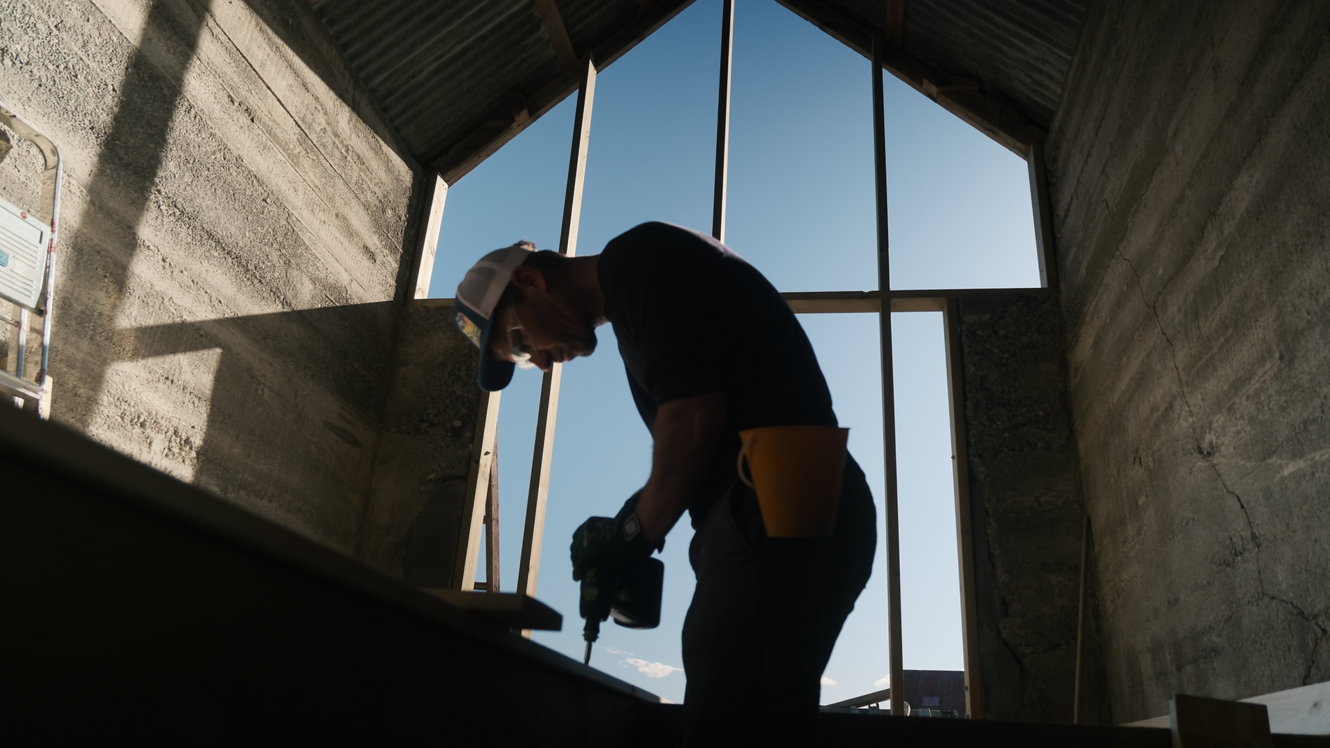 Silhouetted man of Rintala Eggertson Architects using a drill inside a partially constructed building with high ceilings and large windows, conveying industrious focus.