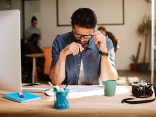 A man sitting at his office desk with a pen in his mouth, staring blankly at his screen, surrounded by paperwork and digital distractions—representing an overstimulated brain and inability to focus.