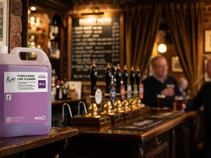 Purple beer line cleaner container on a pub bar counter. Blurred background shows patrons with drinks, brick walls, and a menu board.
