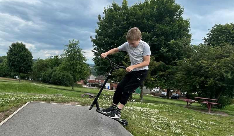 Boy on a scooter doing a trick on a skate ramp in a grassy park under cloudy skies.