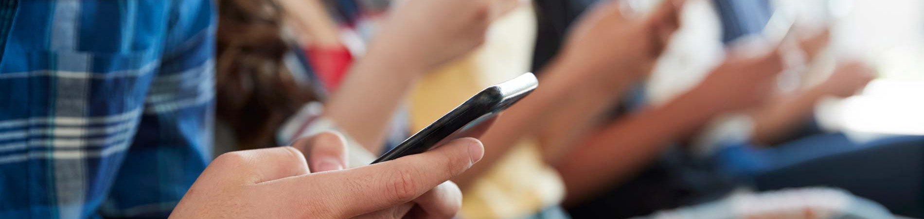 Teenagers seated in a bright room, focused on smartphones, with a casual blue plaid shirt in the foreground
