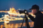 Photographer in a cap captures a night cityscape with a camera on a tripod. Blurry warm city lights illuminate the dark blue sky.