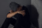 A person wearing a gray knit hat sits against a dark wall, arms crossed over knees, head resting on arms, conveying a somber mood.