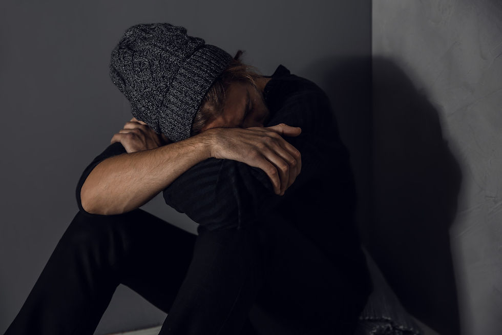 A person wearing a gray knit hat sits against a dark wall, arms crossed over knees, head resting on arms, conveying a somber mood.