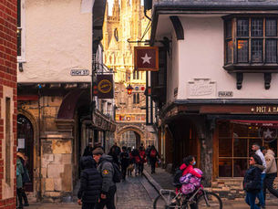 People stroll along a narrow street with shops, historic buildings, and a view of a cathedral in the background. Sign reads "High St."