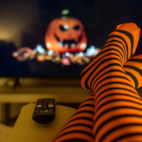 Striped orange-and-black legs crossed on a couch with a remote, watching a spooky pumpkin face on TV in a cozy dark room.