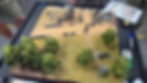 Miniature tabletop battle on a grassy village map with trees, ruined buildings, small figures, dice, and rule sheets around the board