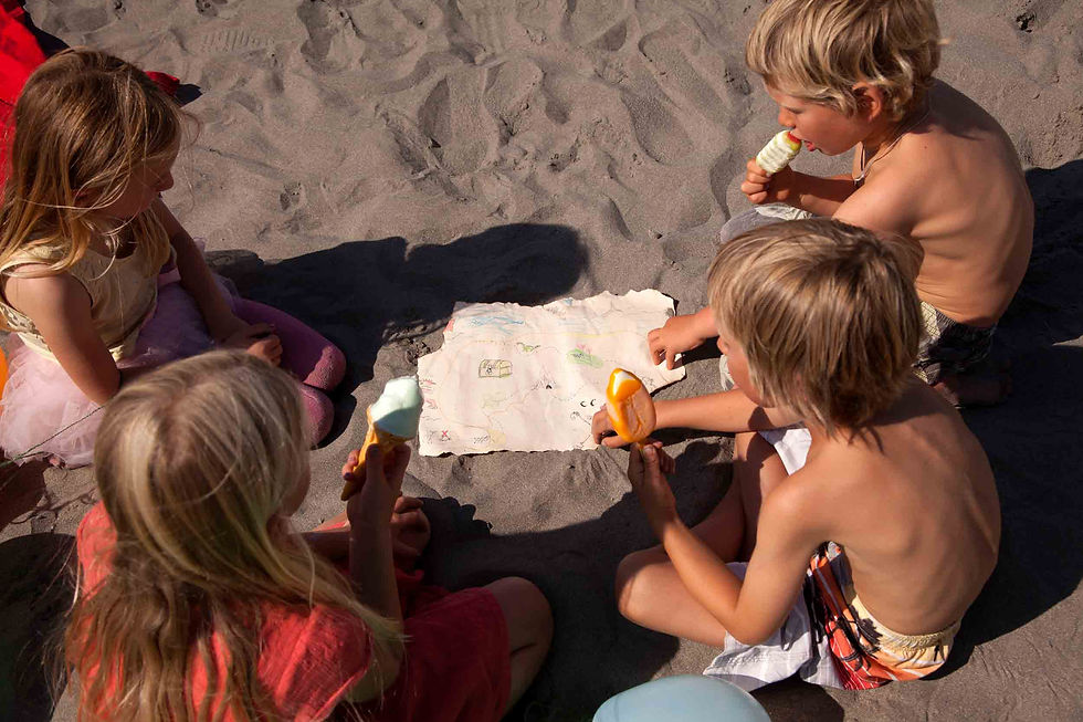 Four children eat ice cream while sitting in a circle on a sunny sandy beach around a paper map drawing.