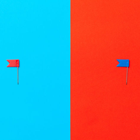 Two small flag-shaped pushpins on a split cyan and red background, one red on blue and one blue on red.