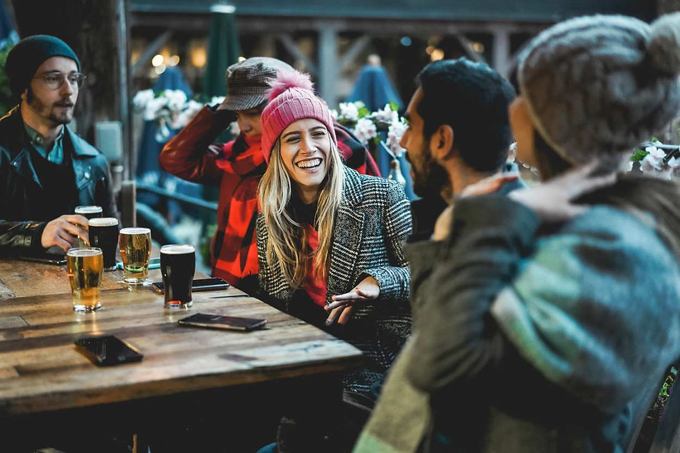Friends laugh and chat at an outdoor table with drinks. One wears a pink hat. Warm, casual setting with wooden table and flower decor.