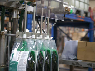 Liquid packaging machine in factory filling green detergent bags. "EVOLUTION" text visible. Cardboard boxes and industrial pipes nearby.