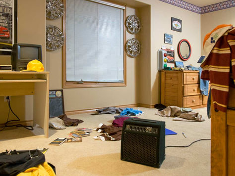 Messy room with clothes on the floor, a TV on a desk, posters, and an amplifier. Walls have metallic decorations, dresser with items. Cozy feel.