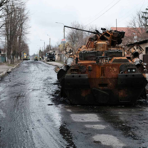 Rusty, destroyed tank on a muddy street lined with bare trees. Distant construction vehicles and workers are visible under a cloudy sky.