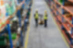 Two warehouse workers in yellow hard hats stand in a long aisle beside a hand truck and stocked metal shelves.