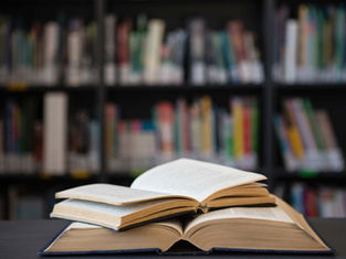 Open books stacked on a table in a library, with blurred bookshelves in the background and a quiet, studious mood.