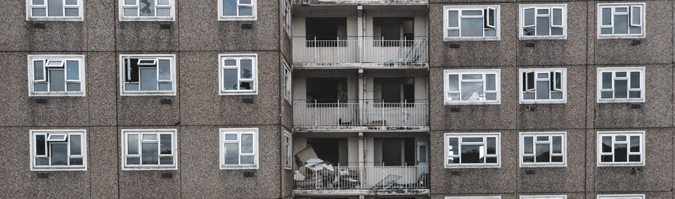 Gray, worn-down apartment building with open windows, broken glass, and debris-filled balconies. Mood of neglect and decay.
