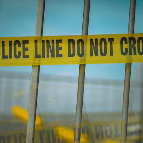 Yellow police tape reading POLICE LINE DO NOT CROSS stretched across metal bars, with a blurred background.