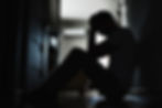 Silhouette of a person sitting on the floor in a dim hallway, head in hands, creating a somber mood. Light filters from a door in the background.