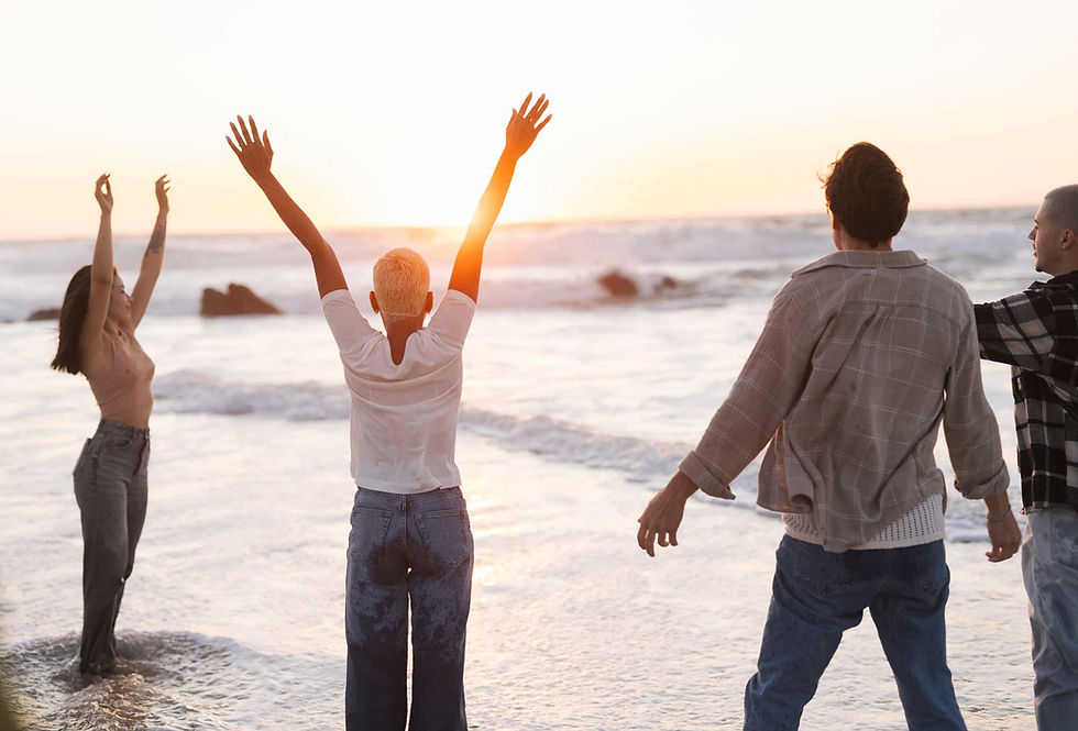 Four people stand on a beach at sunset, arms raised joyfully. They're in casual clothing, surrounded by waves and a glowing horizon.