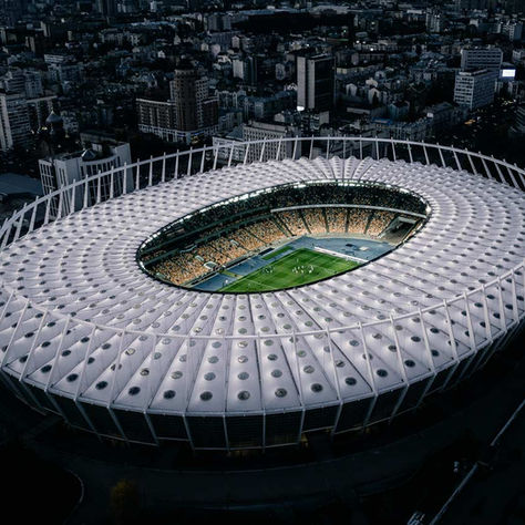 Illuminated stadium at night with a filled crowd, surrounding cityscape in shadow. Bright field center stage, mood is vibrant and dynamic.