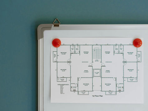 Floor plan pinned to a whiteboard with red magnets, on a blue wall background. Rooms labeled, showing dimensions and layout details.