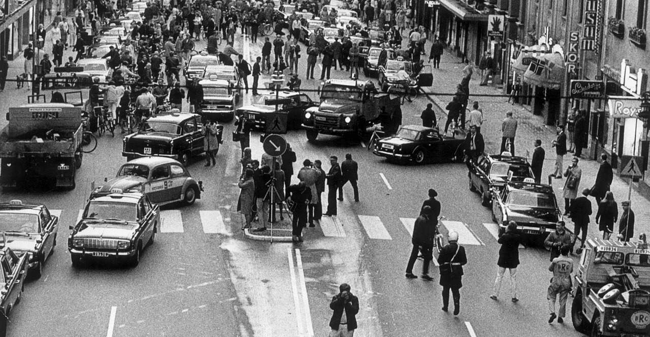 Black-and-white city street crowded with pedestrians and cars, with signs and storefronts lining a tense, busy intersection.
