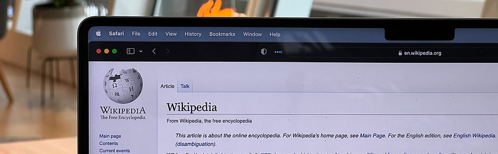 Laptop screen showing the Wikipedia homepage in a web browser. Background is softly blurred with warm tones, suggesting a cozy indoor setting.