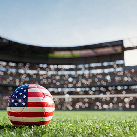 Soccer ball with US flag design on grass field in stadium. Blurred crowd and scoreboard in background. Bright, sunny atmosphere.