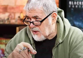 John Blanche with white hair and glasses in a green hoodie speaks thoughtfully indoors, pointing with a ringed hand.