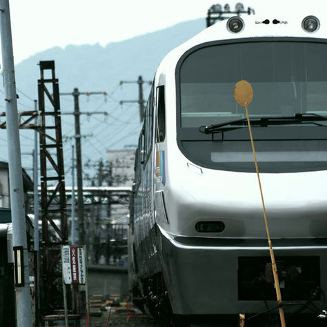 Modern silver train on tracks in an urban area, with mountains in the background. Visible power lines and a sign in the foreground.