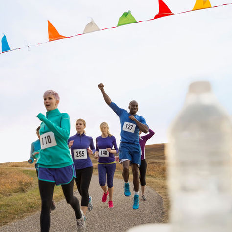 Runners in bright outfits compete on a rural path under colorful bunting. Numbers are visible. Bottles in the foreground. Energetic mood.