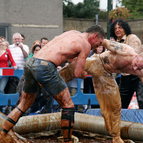 Gravy-covered men wrestle in an outdoor ring while a crowd watches behind blue barriers, creating an intense, chaotic scene.