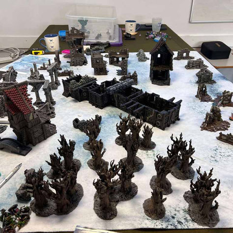 Tabletop game setup with painted miniatures, trees, and buildings on a snowy map. Dice and cups in the background, creating a playful mood.