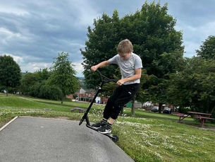 Boy rides a black scooter on a sloped path in a green park, leaning forward under cloudy skies, with trees and picnic tables nearby