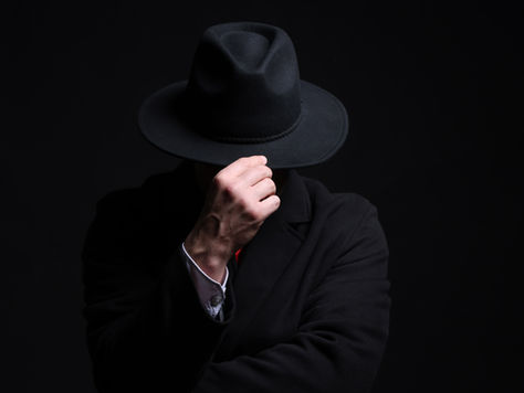 Mysterious man in a black fedora and coat tips his hat, face hidden against a dark black background.