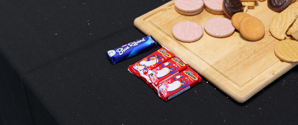 Assorted biscuits on a wooden board beside Blue Riband and Penguin packs on black cloth.