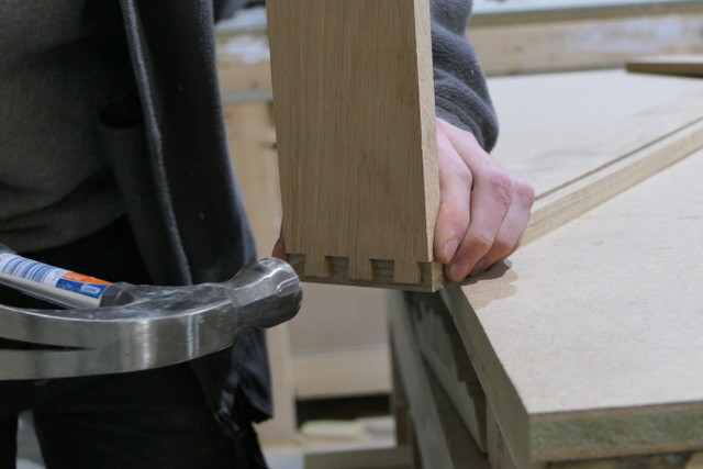 This image has been taken inside our showroom, it show's our cabinetry being made. The person in the photo is holding a hammer and wood. The wood is showing the dovetail joints.